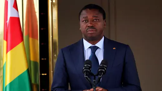 Faure Gnassingbe, president of Togo, wheres a navy suit and dark blue tie as he clasps his hand in front of a double microphone. Behind him and to the side is a Togolese flag hanging on a flag pole.