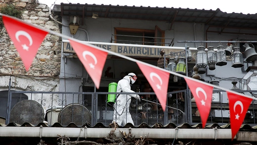 A worker in a hazmat suit sprays disinfectant in Turkey's Grand Bazaar. Turkish flags hang across the image foreground.