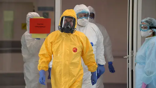 Russian President Vladimir Putin walks toward the camera wearing a yellow protective suit, blue gloves, and a mask.  Other people in protective suits are in the background.