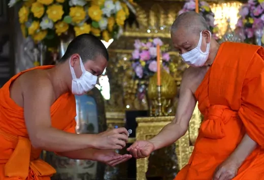A Buddhist monk provides hand sanitizer to another monk. Both wear orange robes and surgical masks.