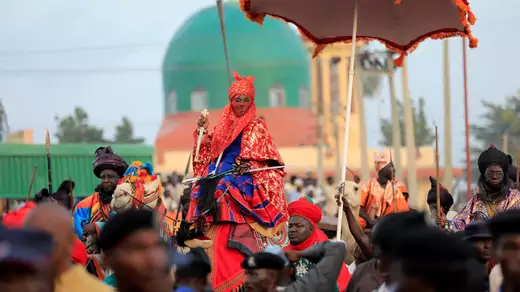 Muhammad Lamid Sanusi II, then the emir of Kano, dress is mostly red robed embroidered with gold and his red turban and veil, rides through a crowd on a dressed camel during the Durbar festival, on the second day of Eid al-Adha celebration in Nigeria's northern city of Kano, on September 2, 2017. An aid holds a heavy red parasol over him as he rides.