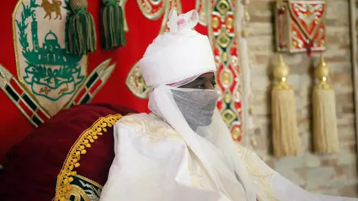 Then-Emir of Kano Lamido Sanusi sits in white clothing with a sheer white vale and white turban, buttressed by a large, ornate, and maroon cushion, flanked by regalia.