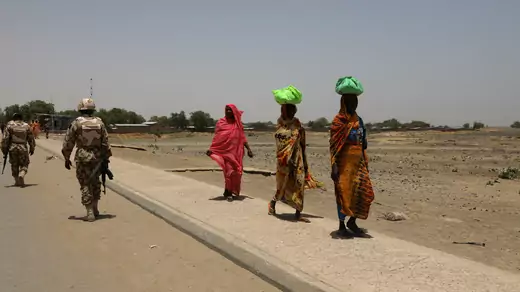 Women, wrapped in red, organge, and blue cloth, carry green parcels on their head and walk past armed soldiers in military fatigues on a bridge separating Cameroon and Nigeria, in Gamboru Ngala, Borno, Nigeria.
