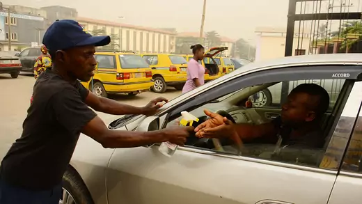 A Nigerian Security and Civil Defense Corps official in a dark shirt and dark blue cap leans on the car of the driver as he sprays the driver's hands hand sanitizer in front of Gbagada General Hospital, in Lagos, Nigeria, on February 14, 2020.