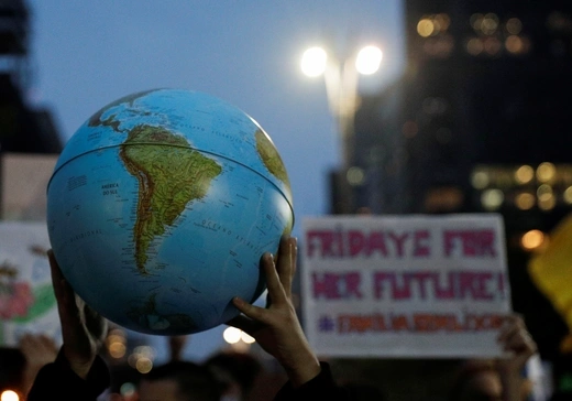Two hands hold a globe in the foreground as climate activists march and display signs in support of the Fridays for Future movement. 