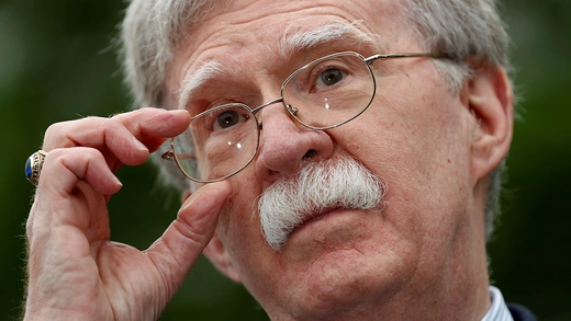A close-up picture of National Security Advisor John Bolton touching his wire-framed glasses and tilting his head.