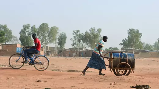 A woman pushes a barrel that resembles an oil drum on its side on a cart over the dusty ground as a man rides the opposite direction on his bike. There are trees and homes in the background. The barrel is filled with water she bought from a privately-owned water tower, amid an outbreak of the coronavirus disease (COVID-19), in Taabtenga district of Ouagadougou, Burkina Faso, on April 3, 2020.