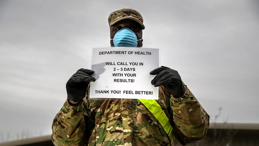 A National Guard service member wears a mask and gloves and holds a sign reading that the Department of Health will call patients within three days with their test results. 