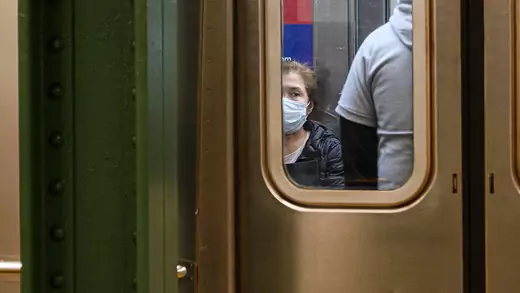 A woman in a mask is seen through the window in a subway car door