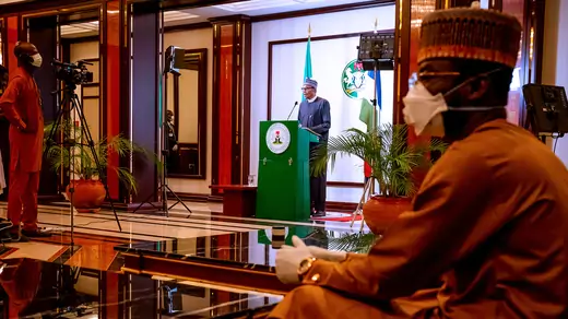 A cameraman with a medical mask films Nigerian President Muhammadu Buhari as addresses the nation on the coronavirus disease (COVID-19), in Abuja, Nigeria. In the foreground is a man sitting with a medical mask and gloves. 