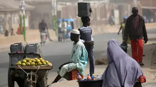 A man sits on a box selling fruit and what look like gasoline bottles from a cart, while a women sells a bottles of something, along a mostly empty road after the postponement of the presidential election in Kano, Nigeria, on February 17, 2019
