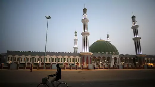 A man cycles past the Al Ansar mosque in Maiduguri. Four red and white minarets are visible around a green dome topped with gold. Person-sized arches line the one-story building around the dome and minarets.