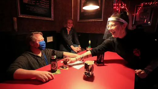 A woman wearing a face guard passes a drink to a man wearing a face mask at a bar.