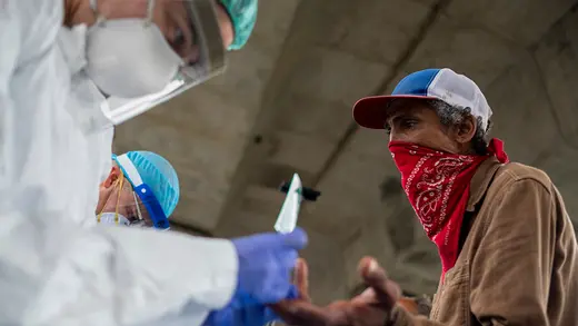 A lab technician wearing a protective suit and gloves and a homeless person wearing a red bandana tied of their face under an overpass.
