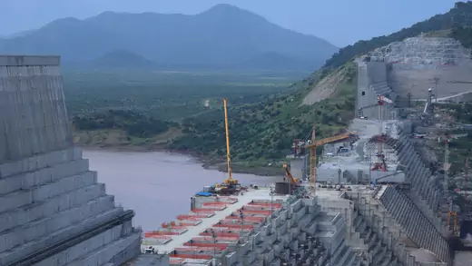 Ethiopia's Grand Renaissance Dam undergoes construction work on the River Nile. The far side of the dam run up against a hewn rockface that rises above the height of the dam. In the distance, a grassy, forested mountain is seen against a dark blue sky.