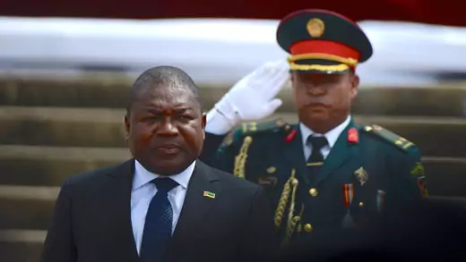 President Filipe Nyusi of Mozambique stands in a dark suit with a blue tie and white shirt as he is sworn in for his second term as president. Behind him, a military officer in ceremonial dress salutes him with a white-gloved hand.