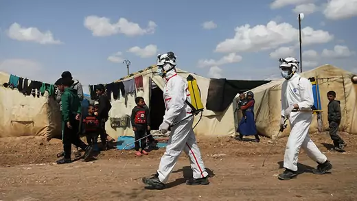 Two members of the Syrian Civil defense in protective gear sanitize a internally displaced persons camp with several children in Syria