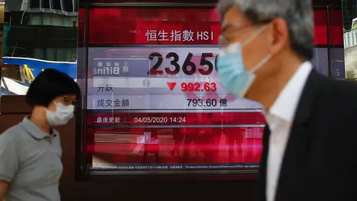 Two people wearing masks cross a panel display of the Hang Seng Index during afternoon trading in Hong Kong, China