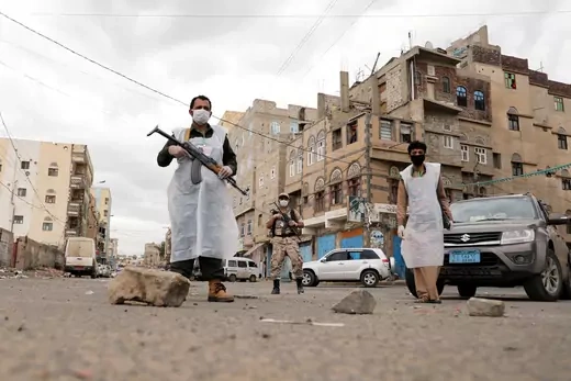 Three men wearing protective clothing and masks--two of whom have guns--stand guard in front of cars parked in the middle of a debris-ridden street during a twenty-four hour curfew in Sanaa, Yemen, on May 6, 2020. 