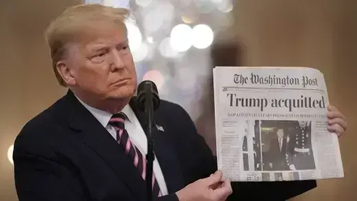Dressed in a suit and standing in front of a microphone, Trump holds up a copy of the Washington Post showing news of his acquittal.