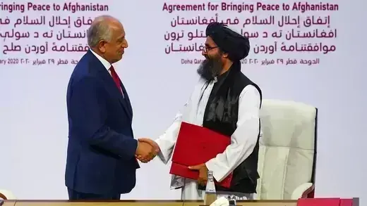 A photo of two men shaking hands in front of a screen that reads "Agreement for Bringing Peace to Afghanistan."  The man on the left is dressed in a navy-colored suit, and the man on the right has a beard, is wearing a head covering, and is carrying a red folder.