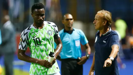 Nigeria's John Obi Mikel, with a water bottle, drenched in sweat in a green, white, and black patterned Nigeria jersey wears number 10. He is with coach Gernot Rohr at the Africa Cup of Nations in June 2019. A referee in a blue top and black shorts is in the background.
