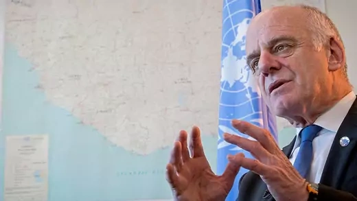 Dr. David Nabarro at the United Nations Headquarters with a blue UN flag and a large map behind him.