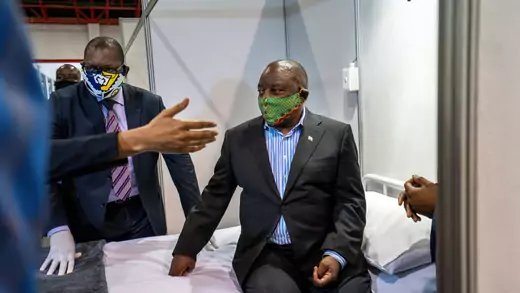 South Africa President Cyril Ramaphosa, in a dark grey suit without a tie and a green facemask, sits on a made bed at a coronvirus treatment center. A hand gestures in the foreground and a man in a suit and tied with a mask flanks Ramaphosa. 