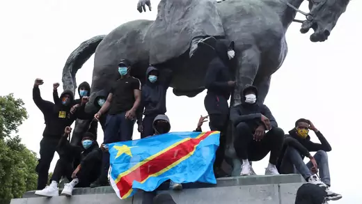Protestors dressed mostly in black with masks and face coverings display the DRC flag on the pedestal of a massive statue of King Leopold II on a horse. The sky is white and there is green foliage from a tree in the back left of the photo.