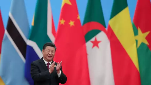 President Xi Jinping of China claps and smiles in a dark gray suit with a maroon tie and white shirt. Behind him are national flags on vertical flag polls. The Chinese flag is front and center. It is flanked by the flags of South Africa and Botswana to the left, and of Algeria, Benin, and Burkina Faso to the right.