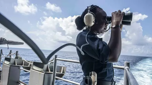 A woman on a naval vessel looks at the sea through binoculars. 
