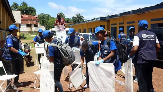 WHO workers in dark blue vests and light blue hats arrange chairs between two rows of buildings as they set up a vaccination center for Ebola in the Congolese port city of Mbandaka in 2018. 