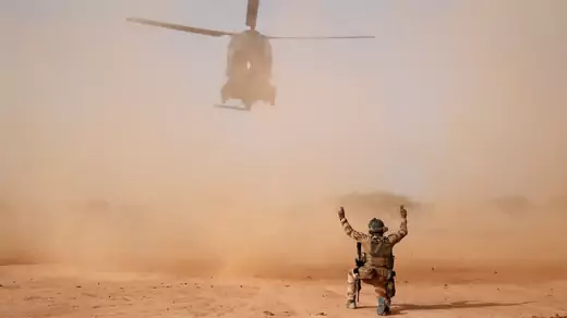 Dust is whooshed into the air as a military helicopter prepares to land. A soldier in desert camouflage kneels on one knee as he signals the helicopter. The sky, obstructed by the dust, is otherwise a dull, clear blue. 