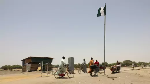 People push carts and bicycles near a flag pole with the Nigerian flag as they begin to cross a road bridge. The ground is dry and dusty, the cloudless sky is dull grey-blue, and small wooden structure is in the background. The canopies of green trees can be seen in the distance. 