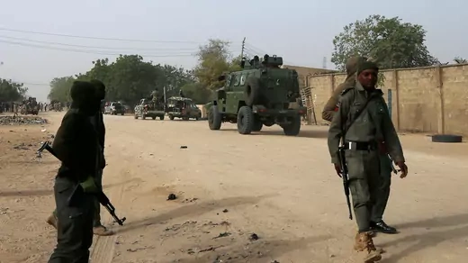 Three soldiers walk on a dirt road with rifles. In the background, armored troop carriers drives on the dirt road along a wall with trees further down the road. 