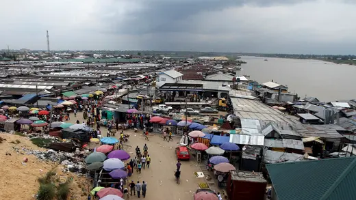 A picture of a road running through Swali market, likely taken from an overlook. The road is lined with solid-colored, large umbrellas. People are on the road. The market appears to be made of small shacks mostly creating one large, single-story, structure. In the distance to the right is the river. The sky is gray. 