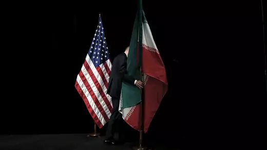 U.S. and Iranian flag on a stage with a black backdrop while a staff member is removing the Iranian flag off the stage