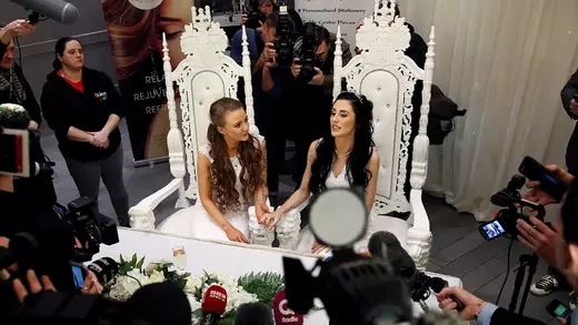 Two young women sitting in ornate white chairs and holding hands surrounded by members of the media.