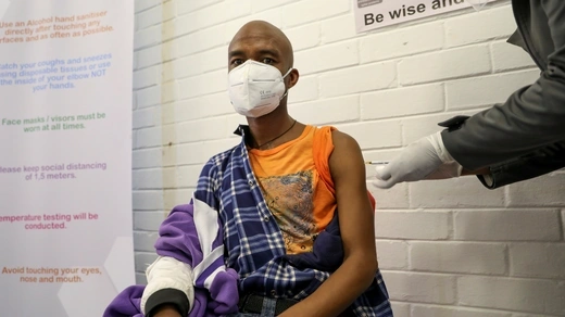 A bald, young man with a white mask sits, with three layers of clothing, a purple sweater, a dark blue flannel, and finally, an orange t-shirt, peeled back in order to reveal his harm as he receives a vaccine candidate for COVID-19. The health care worker administering the vaccine is in a dark grey jacket and is wearing white medical gloves. The wall is painted white brick and there are informational posters around. 
