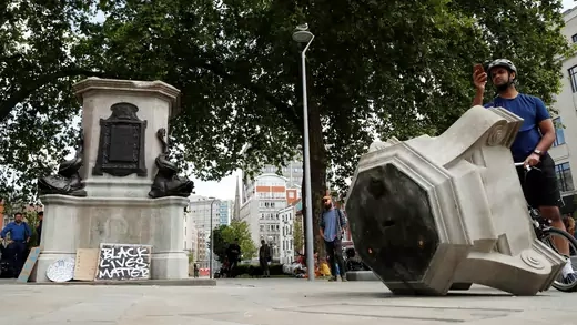 A man wearing a bike helmet and holding his bike takes pictures with his phone of the pedestal that formerly held a statue of Edward Colston. In the background is the structure on which the statue was held, with bronze statues and with a black lives matter sign visible. 