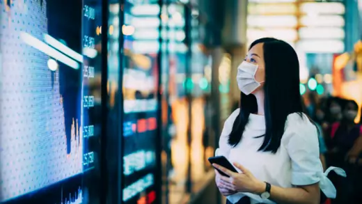 Businesswoman wears face mask and checks financial trading data by the stock exchange market display screen board.