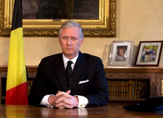 Belgian King Philippe sits at a dark wooden table in a black suit, black tie, and white shirt. His hands are folded and his face is somber, with his silver-white hair combed down. To the left is a Belgian flag on a vertical flag pole; to the right are two family framed pictures sitting on a low bookcase.