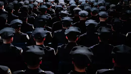 A photo from behind of rows of police officers wearing navy blue uniforms and hats in a moodily lit arena.