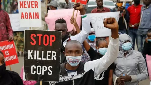 I young black man with a light blue medical mask on which is written in red "I can't breathe," looks in the direction of the camera, with his right hand holding a black sign that says "Stop Killer Cops" and his left hand raised in a fist. He is flanked by other protestors in masks and with signs. 