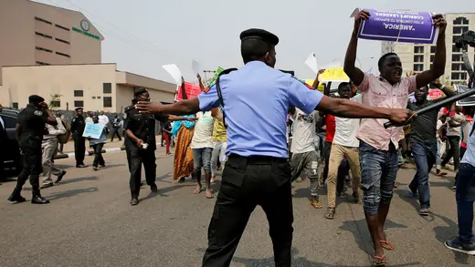 A police officer in dark pants and a blue, short-sleeved shirt with a black cap, tried to block the path of protestors carrying signs on a street in Abuja. He is armed with a baton. 