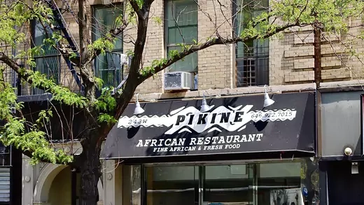A black restaurant awning is pictured that says "Pikine" and "African Restauran" and "Fine African & Fresh Food." The restaurant is on the first floor of a light brick apartment building. A tree with thin foliage is to the left of the restaurant.