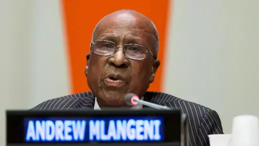 Andrew Mlageni, an aged, bald man with glasses, sits at a dais. His name is on a lit-up placard in front of him, as is a thin microphone. He is speaking.