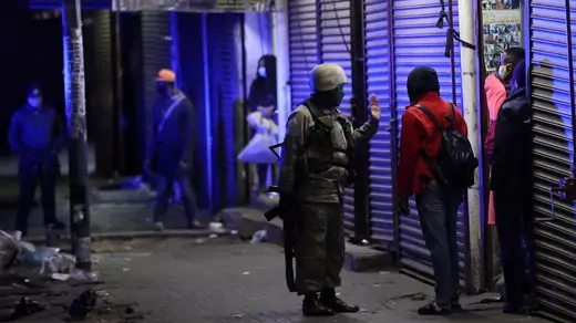 A soldier in military fatigues holds his rifle in one hand while he gestures to a man in a red coat with a backpack. They are standing on a street of shops with metal gates pulled down, lit by blue-purple light. Everyone in the picture is wearing masks. 