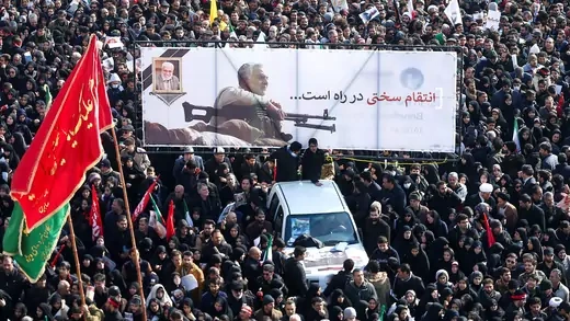 A crowd surrounds a car carrying a banner depicting Iranian Quds Force commander Qasem Soleimani