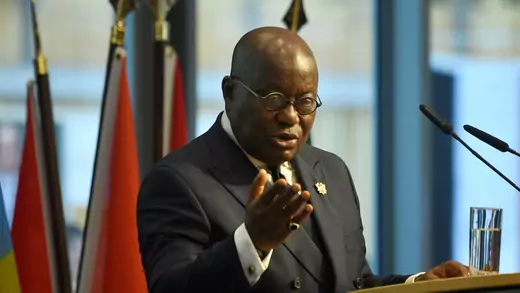 The president of Ghana, a bald African man in a dark suit with glasses, gestures as he speaks at a podium. He is flanked by national flags. 